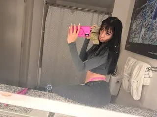 realcookiebangz poses in a grey long-sleeve crop top and matching grey pants, looking at her reflection in a mirror with her long dark hair down.