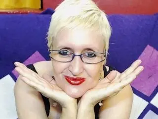 Smiling blonde woman with glasses and red lipstick resting her chin on her hands in a cozy purple setting.