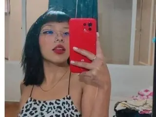 rose_sexxx poses in a black and white spotted tank top, looking at the camera with red lipstick and glasses.