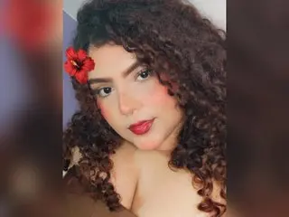 roxxxana19 poses with curly hair and a red flower behind her ear, showing her face and shoulders against a soft background.