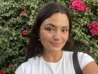 safiya_georgette smiles while looking at the camera, wearing a white t-shirt with her long dark hair down against a background of pink flowers and green leaves.