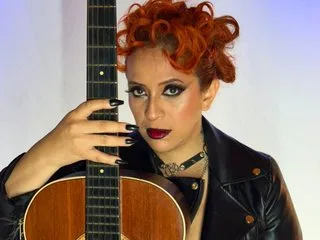 sahratayl0r poses with a guitar, wearing a black leather jacket and a silver choker with dark eye makeup and red lipstick.