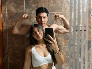 scarlataandluca poses shirtless in a white tank top, flexing her toned arms and showing off her fit physique against a brown tiled wall.