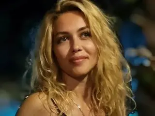 sellenya with long blonde wavy hair, looking at the camera with a slight smile in a dark setting.
