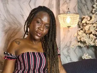 sexy_broown poses with her long braids, wearing a red and black striped off-the-shoulder top against a marble wall background.