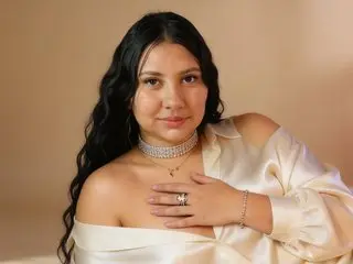 sexy_ssofia poses with her long dark hair down, wearing a white satin off-the-shoulder top against a plain tan background.