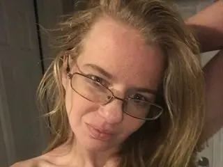 sexysandy99 looks at the camera with blonde hair and glasses, showing her bare shoulders and chest in a close-up shot.