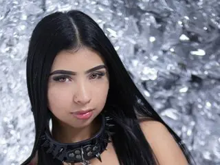 Young woman with long black hair wearing a black studded choker against a shimmering silver tinsel background.