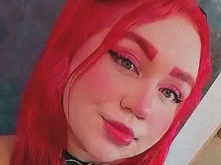 Smiling woman with vibrant red hair and pink makeup wearing a black choker in a soft indoor setting.