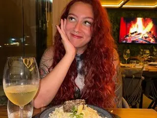 spicyspiders21 with long red hair poses with a hand to her face while sitting at a table with a glass of white wine.