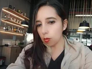 sublimesquirt18 looks at the camera with long dark hair and red lipstick, wearing a light tan jacket over a black top.