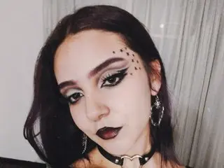 Woman with gothic makeup, dark lipstick, and star face gems wearing a black choker in a moody indoor setting.