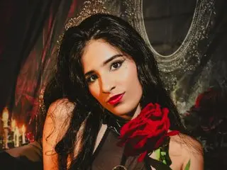 tatianacrow poses with long dark hair and red lipstick, wearing a red rose near her chest in a dark setting.