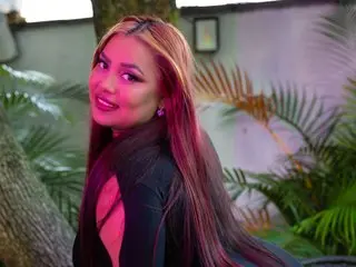 tatikaanyxx smiles while posing outdoors with long reddish-brown hair and a black top near tropical palm leaves.