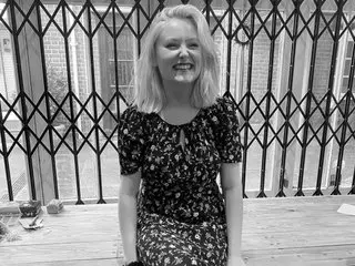 thegoddessvenus is a smiling blonde woman wearing a dark floral print short-sleeve top and matching skirt, sitting outdoors in front of a metal gate.