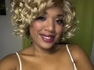 thekelliecapri smiles with her blonde curly hair down, wearing a white strap top against a green background.