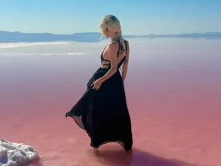 theselenarainx poses in a long black dress with a low back, standing in a pink salt flat setting with her blonde hair down.