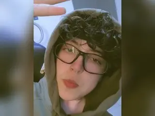 Young man with curly hair and glasses wearing a grey hoodie in a casual indoor setting.