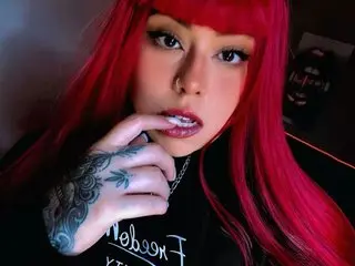 Woman with vibrant red hair and tattoos wearing a black graphic tee in a moody indoor setting.