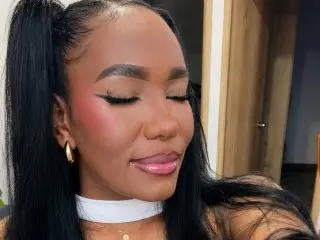 Black woman with sleek high ponytail and glamorous pink makeup wearing a white choker in a bright indoor setting.