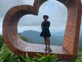 willowsweett stands in a large wooden heart frame overlooking a lush, misty mountain landscape while wearing a short black dress.