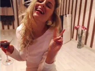 Cheerful blonde woman in a white long-sleeve top holding a cocktail and making a peace sign in a cozy room.