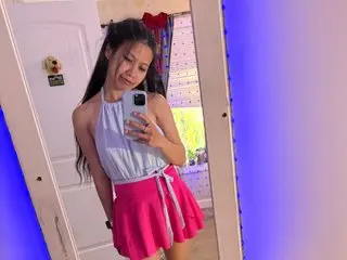 xXxJuicyPinaypplexXx poses in a white tank top and pink skirt, looking at the camera while holding her phone for a mirror selfie.