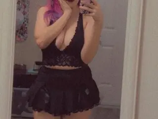 Young woman with pink hair in a black lace crop top and ruffled mini skirt posing in a bedroom mirror.