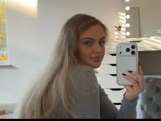 xhaileyrae poses for a mirror selfie, showing her long blonde hair and a grey long-sleeve top against a bright indoor background.
