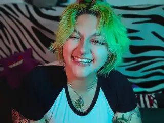 xxgreengoodxx smiles with short green hair, wearing a black and white t-shirt while sitting in front of a zebra-print background.