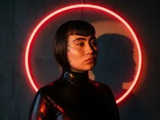 zoee77 poses in a black latex turtleneck, looking forward with short black hair against a dark background with a red neon circle behind her.