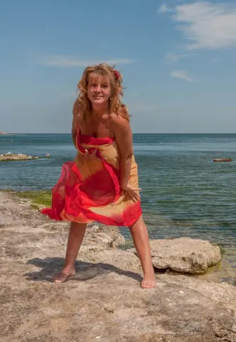 Smiling woman in a flowing red and orange summer dress standing on a sunny rocky beach by the blue ocean.