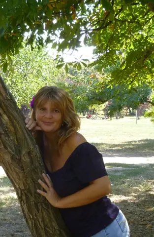 Smiling blonde woman in a navy off-the-shoulder top leaning against a tree in a sunny summer park.