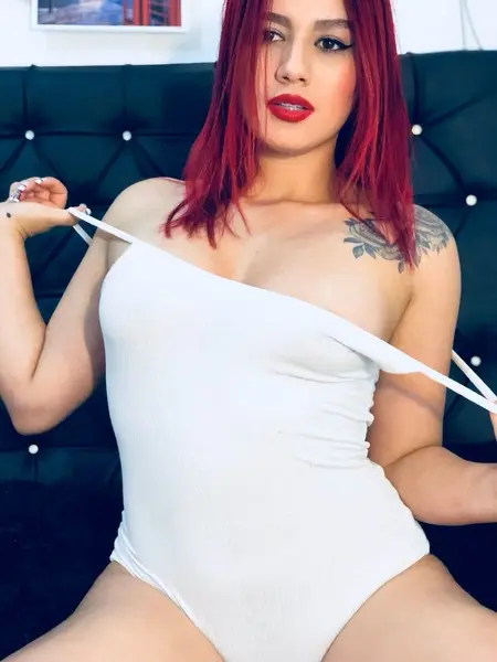 AishaCollins poses with her hands near her face, wearing black fishnet gloves and showing her long orange hair and red lipstick.
