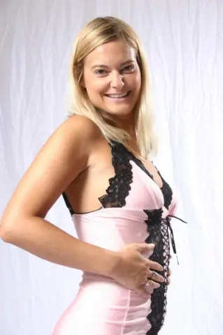 Smiling blonde woman in a pink satin and black lace slip dress posing against a white background.