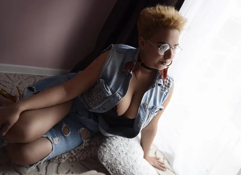 AllyiahH poses in a light tan jacket and red top, looking down while sitting on a white bed frame.