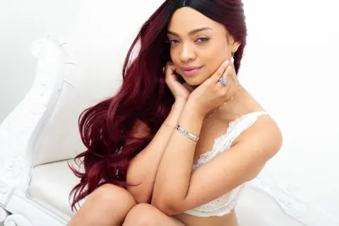 Smiling woman with long burgundy hair wearing a white lace top in a bright, minimalist setting.