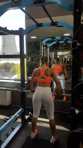 Athletic woman in orange sports bra and white leggings training with a barbell in a bright gym.