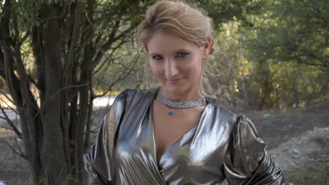 AshleySweetXxX wearing a shiny silver dress and posing outdoors in front of trees.