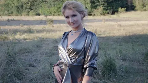 AshleySweetXxX poses in a shiny silver dress while standing in a grassy field.