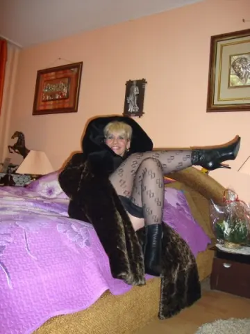 Smiling woman in a dark fur coat and patterned tights relaxing on a purple bed in a cozy bedroom.