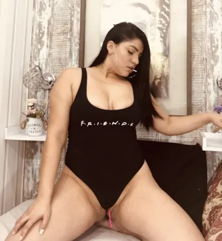 Curvy woman in a black sleeveless bodysuit relaxing in a bright, modern bedroom setting.
