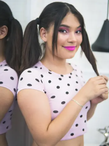 Young woman with dark pigtails and pink makeup wearing a white polka dot crop top in a bright indoor setting.
