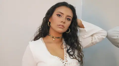 Young woman with wavy hair wearing a white blouse and leopard print choker in a minimalist indoor setting.