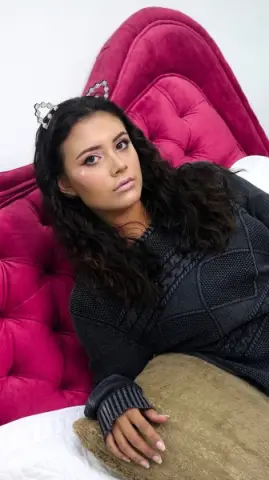 Young woman in a dark grey sweater relaxing on a pink velvet headboard with a cozy, moody aesthetic.