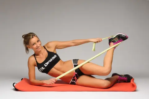 Smiling fit woman in black sports bra doing leg stretches with a resistance band on a red yoga mat.