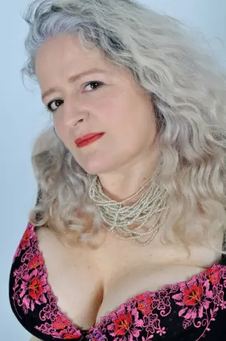 Elegant woman with wavy grey hair wearing a pink floral lace top and layered pearl necklaces.