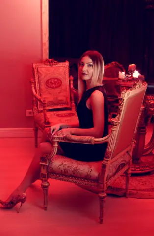 Elegant woman in a black dress sitting in a vintage armchair within a moody, red-lit luxury room.