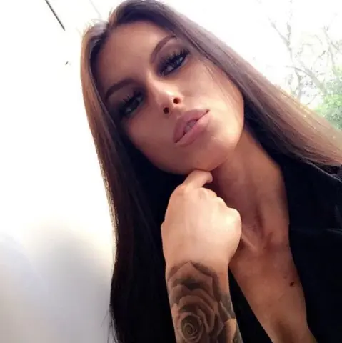 Inkedgirluk poses with her hand on her chin, showing off her long brown hair and large rose tattoo on her arm.