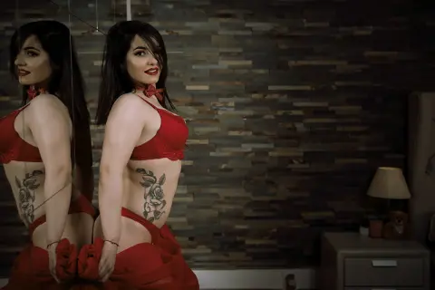 Confident woman in red lingerie and matching skirt posing in a moody, modern bedroom setting.